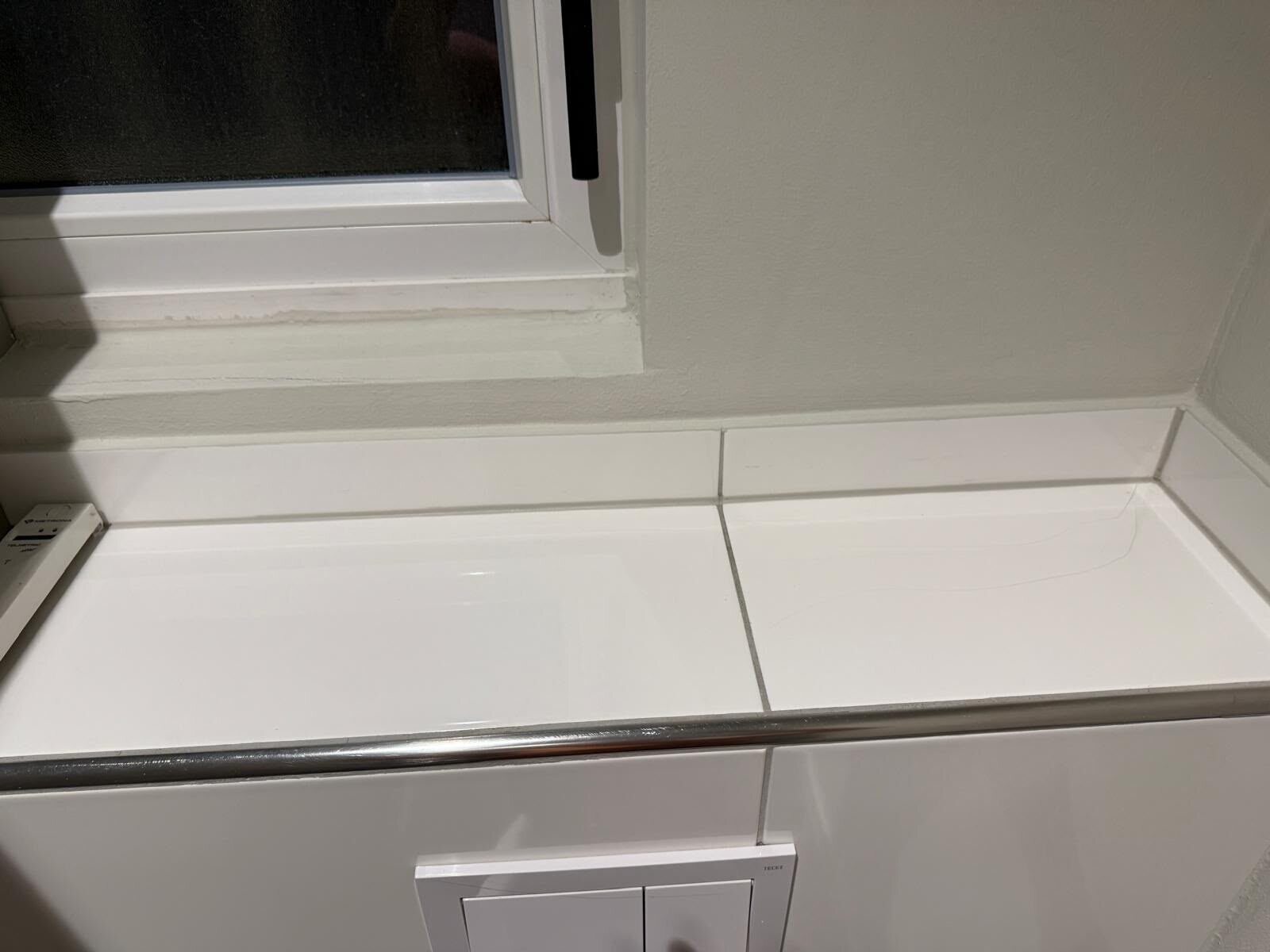 Weiß geflieste Ablage unter einem Fenster mit einzelnen Rückständen