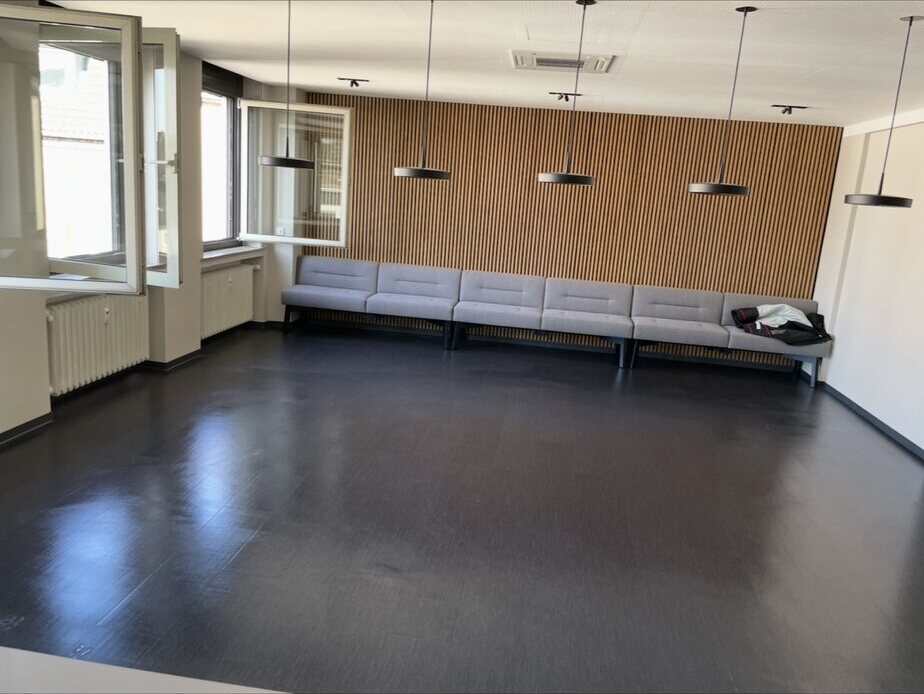 Dunkler Boden mit Lichtreflexionen vor grauer Sitzbank und Holzlamellenwand