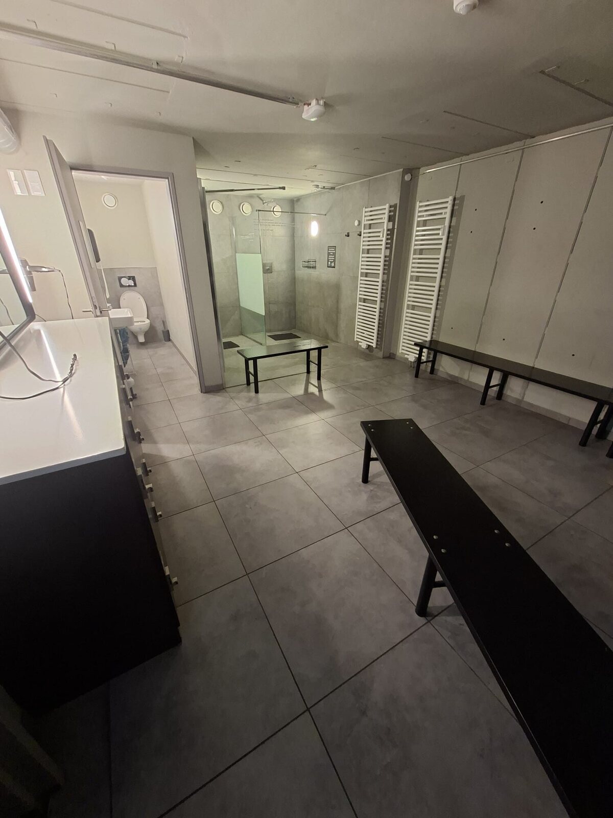 Umkleide mit grauem Fliesenboden, schwarzen Sitzbänken und offenem Zugang zum WC