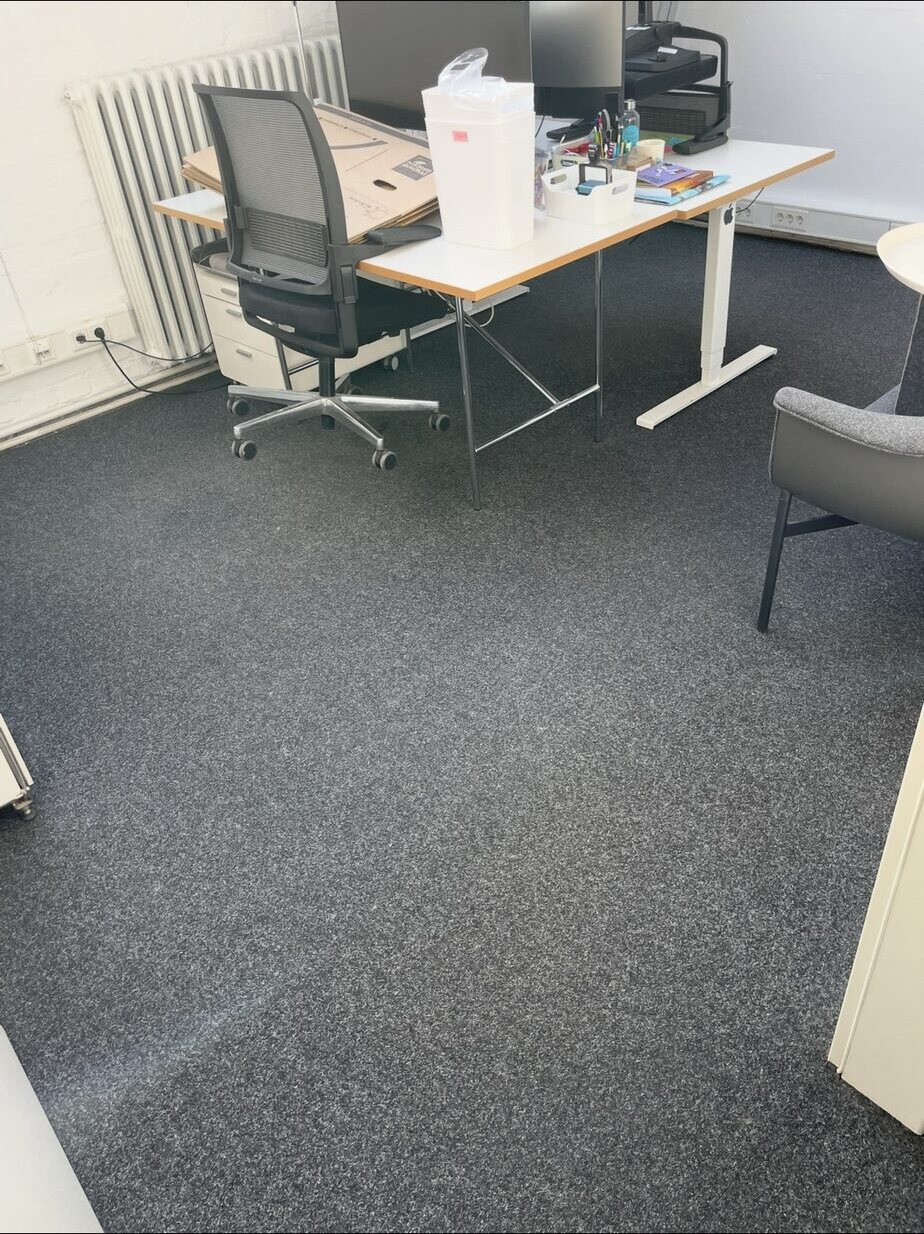 Grau melierter Teppichboden rund um einen mit Büromaterial belegten Schreibtisch