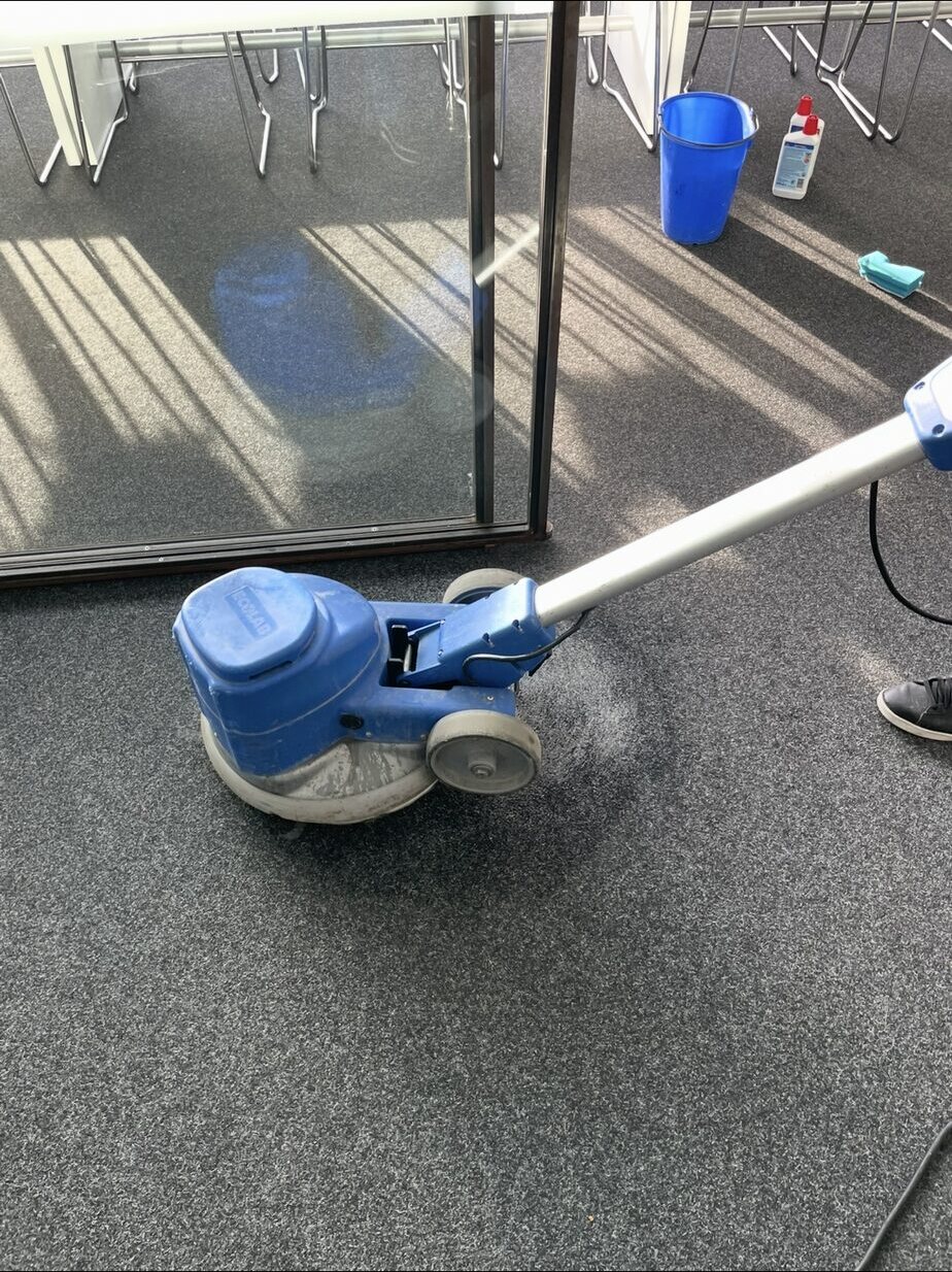 Blaue Einscheibenmaschine mit Gebrauchsspuren auf meliertem Teppichboden neben einer Glastrennwand