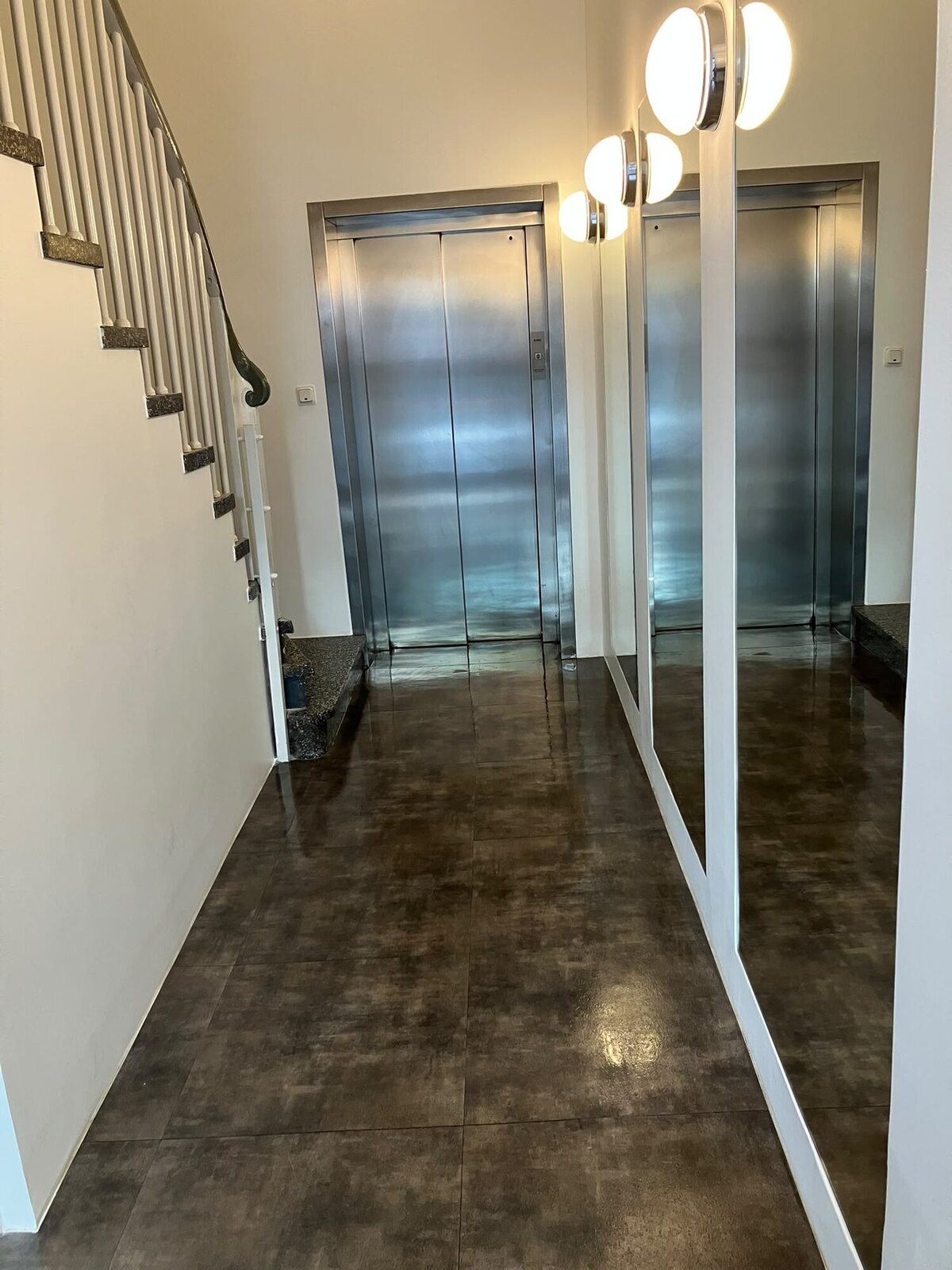 Treppenhaus mit glänzendem Fliesenboden, metallenen Aufzugtüren und verspiegelter Wand