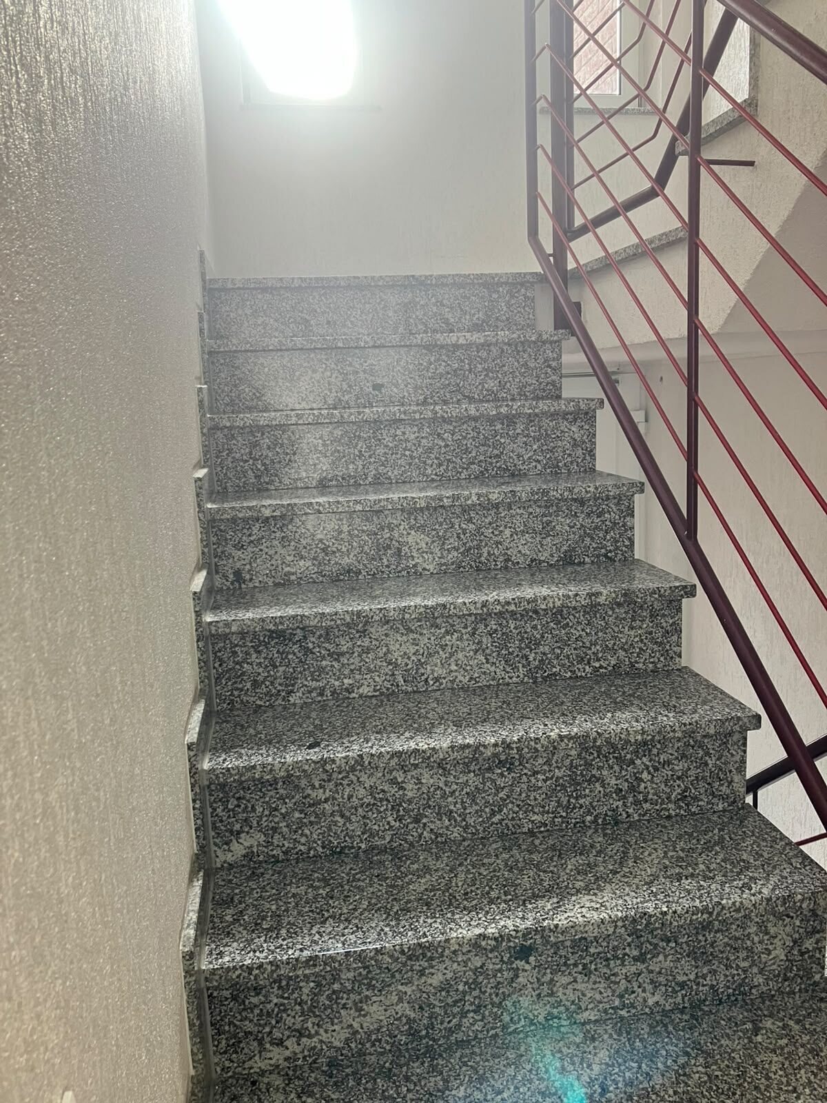 Treppenhaus mit grau gesprenkelten, schmutzfreien Stufen und dunkelrotem Metallgeländer